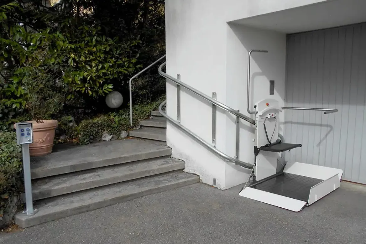 A Savaria inclined platform lift at the foot of exterior steps, its platform unfolded with the ramp flat on the paving and its rail running up alongside the stairs